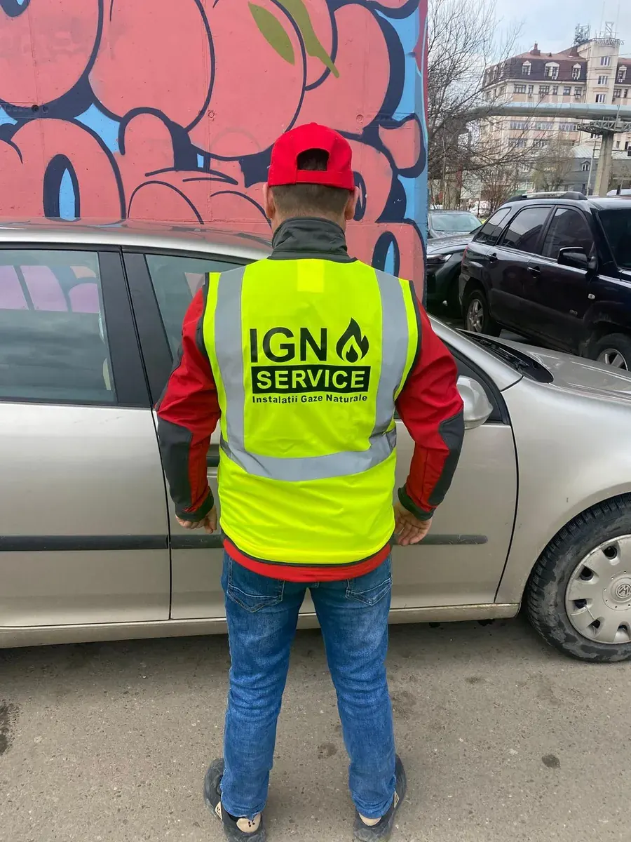 Instalator IGN Service din spate, în vestă reflectorizantă și șapcă roșie, pe trotuar între mașini
