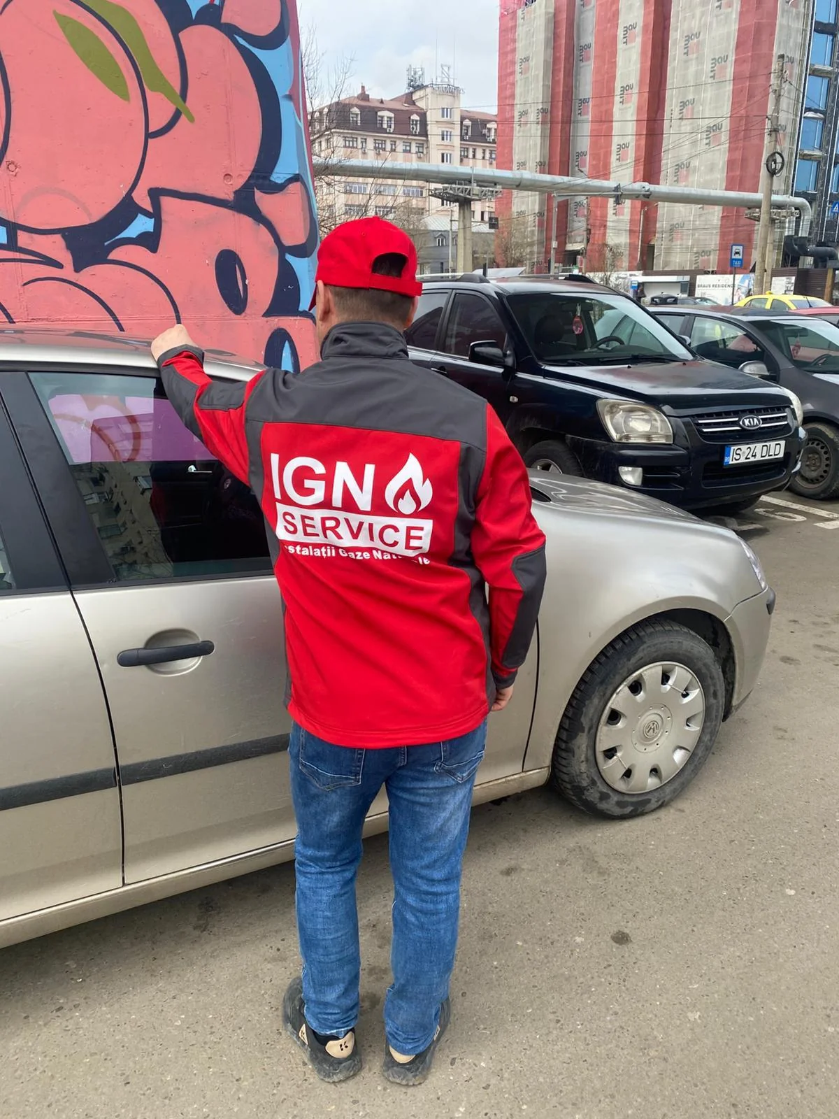 Tehnician IGN Service în geacă roșie și șapcă, văzut din spate lângă mașină pe stradă, în Botoșani